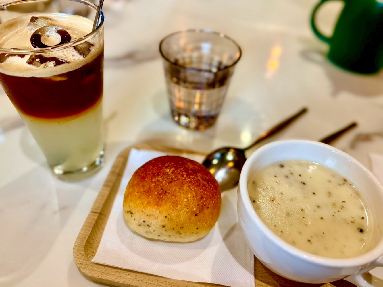 圖片中有一杯分層的冰飲、玻璃杯、麵包及一碗奶油玉米濃湯，擺放在白色桌面上，看起來是一份簡單的餐點。