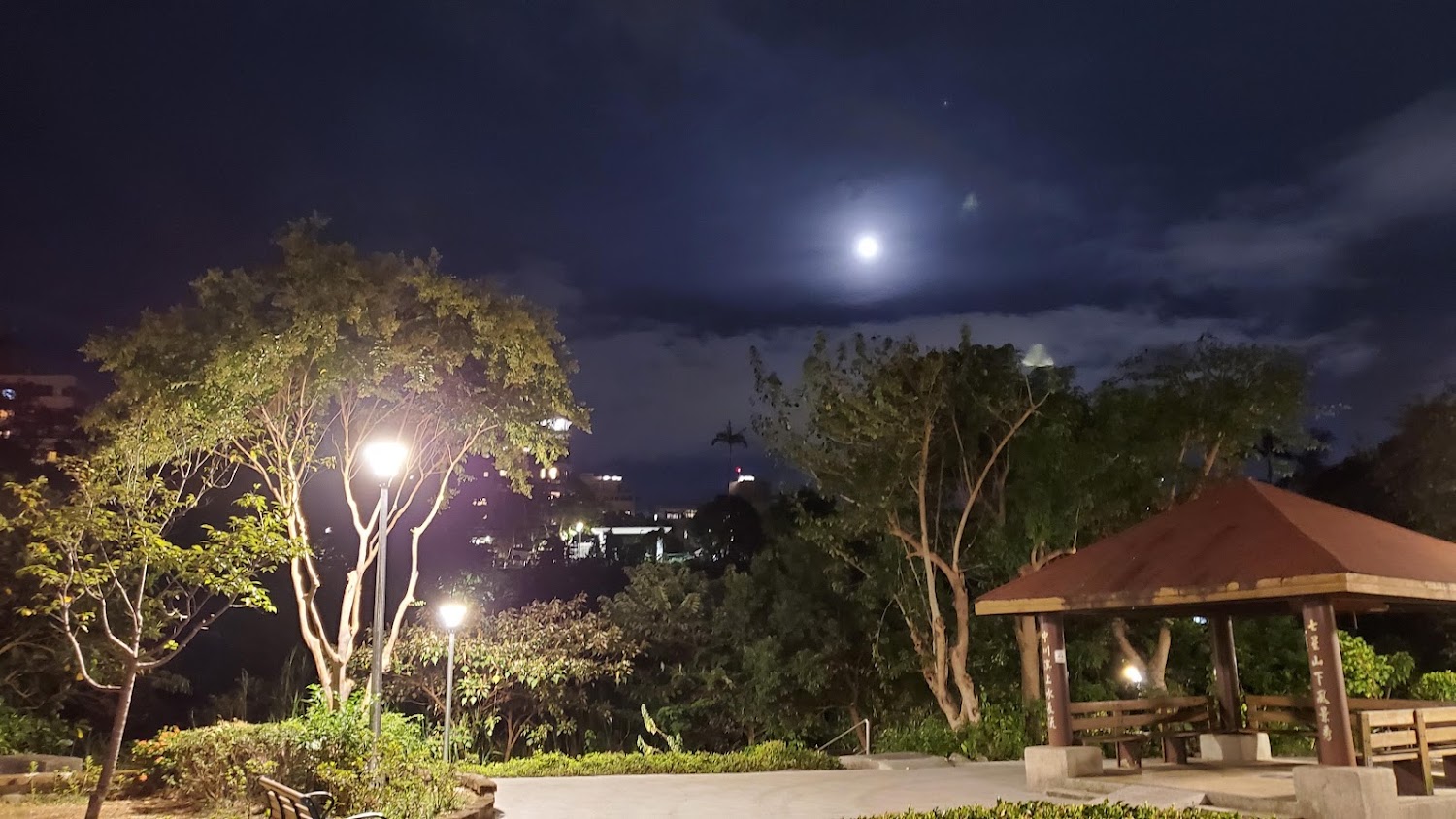 夜晚的公園有樹木、長椅、路燈和有頂棚的涼亭，天空可見月亮