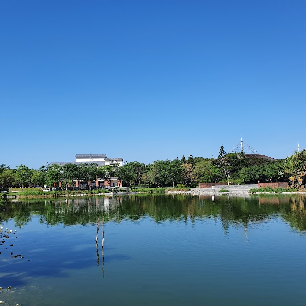 這張圖片展示了一個湖泊，湖邊有綠樹和建築，藍天無雲，景色寧靜。湖面反射出樹與建築的倒影，環境自然且悠閒。