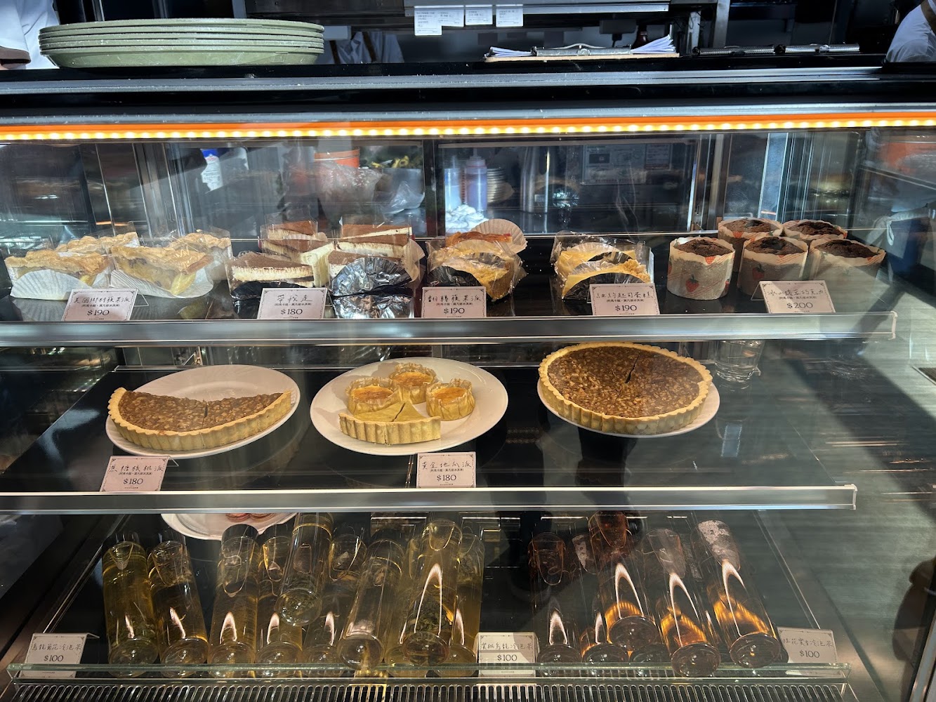 這張圖片顯示一家甜點店的展示櫃，裡面有多種蛋糕、塔類和布丁甜點，標示著不同價格。下層還有透明玻璃杯裝的甜品，整體看起來非常豐富精緻。