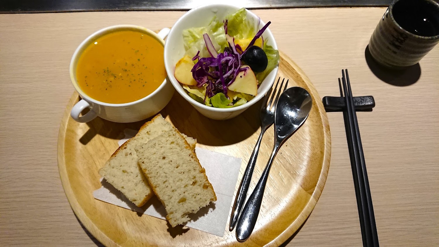 這張圖片主要有一份套餐，包括一碗南瓜湯、一碗生菜水果沙拉、兩片麵包，以及湯匙、叉子和筷子，擺放在木盤上。