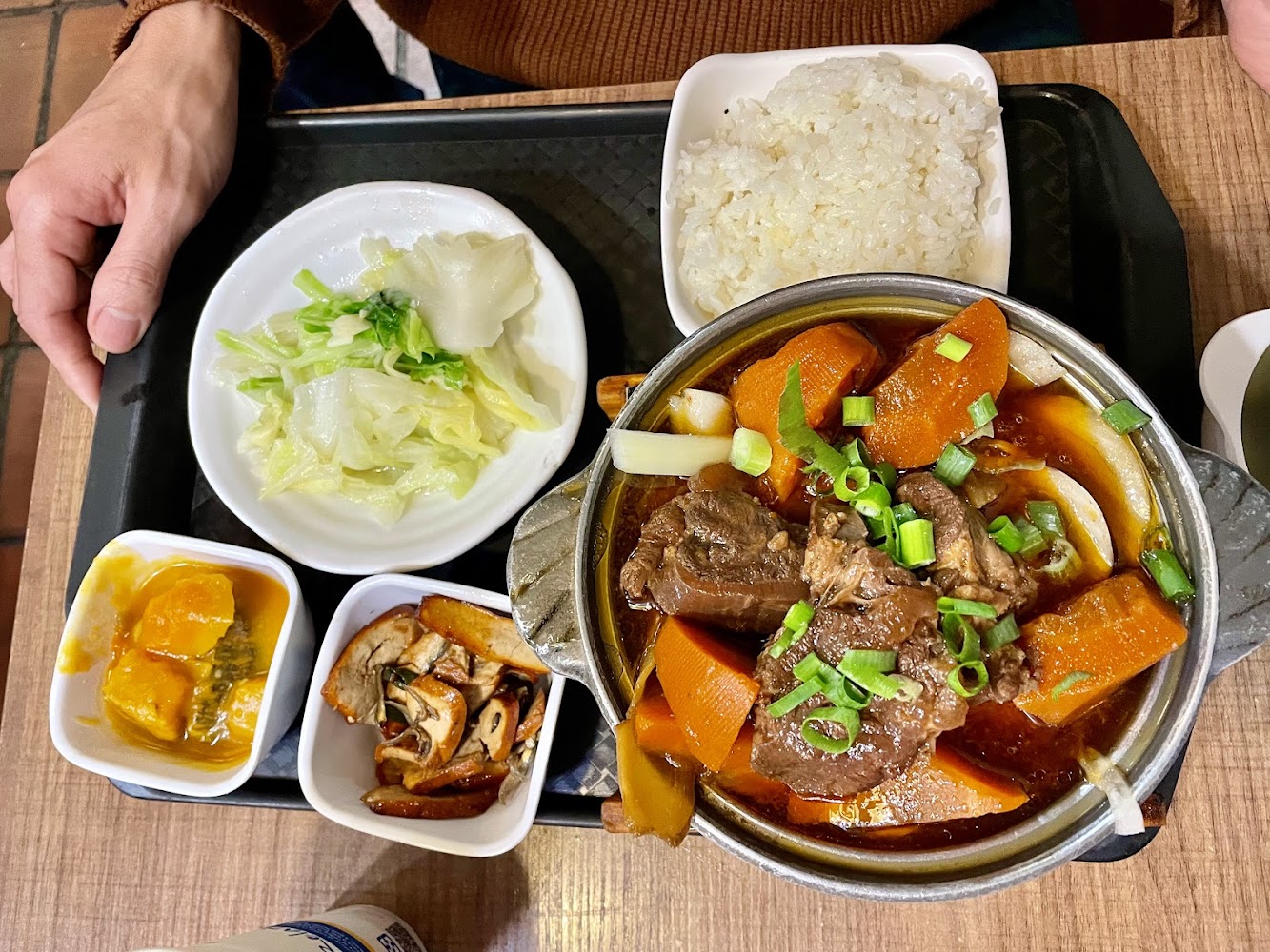 這張圖片展示了一份中式套餐，主菜是紅燒牛肉鍋，搭配白飯、燙青菜、滷南瓜和炒菇類蔬菜，色香味俱全。