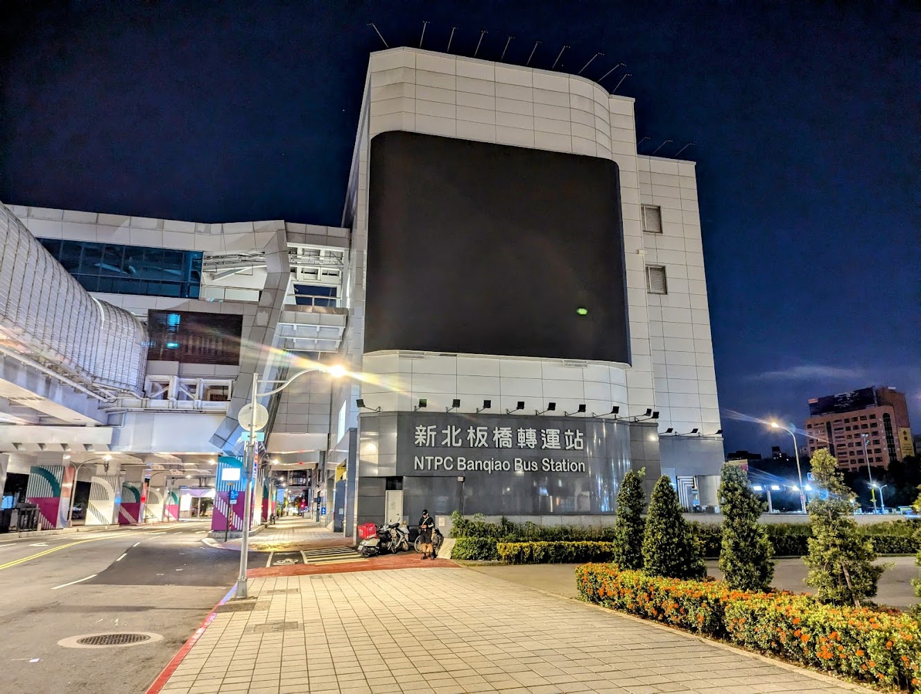 新北板橋轉運站外觀，夜晚燈光明亮，建築現代化，周圍有綠化植栽及摩托車停放