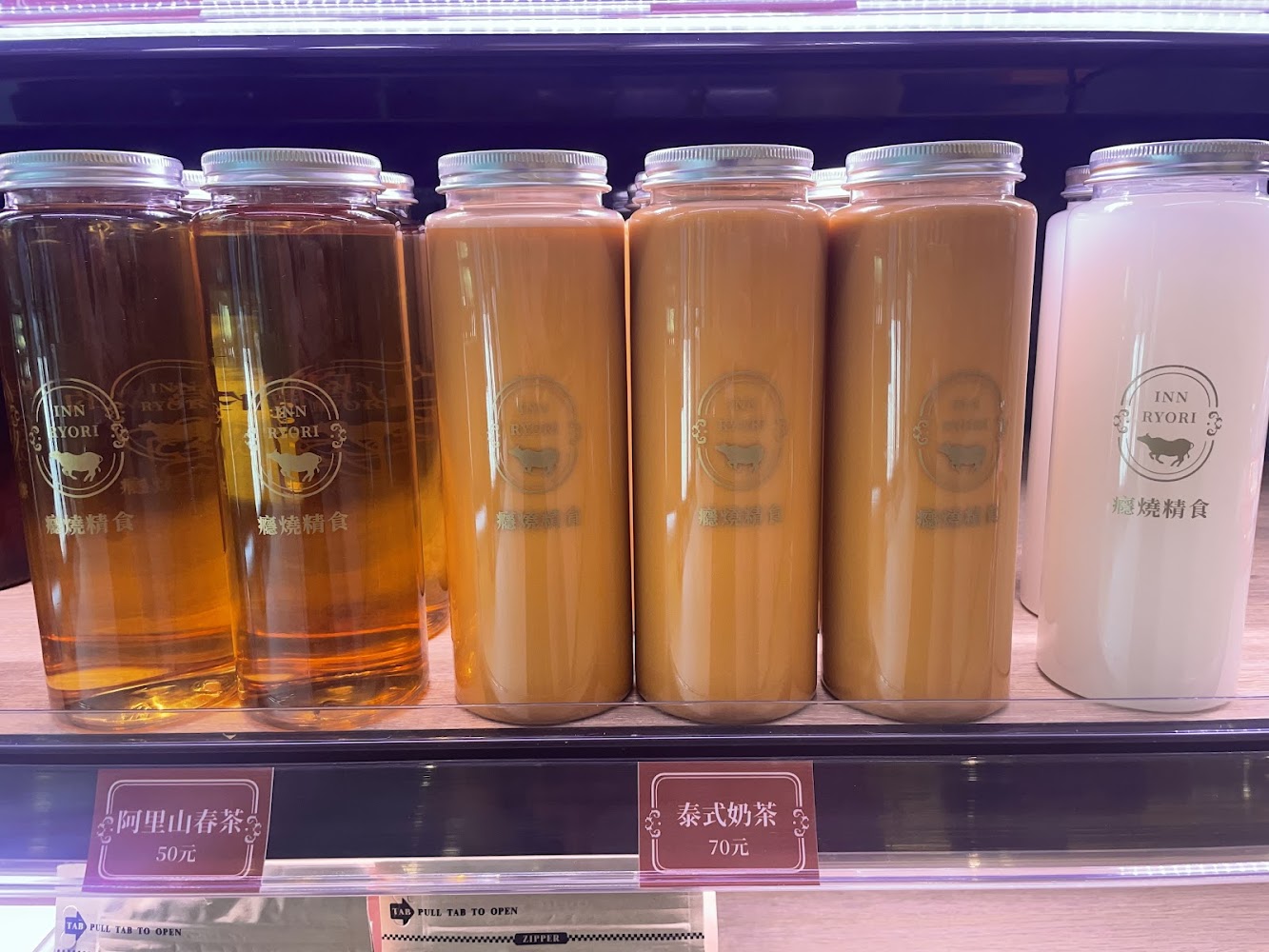 這張圖片展示了三種瓶裝飲品，分別是阿里山春茶、泰式奶茶和另一種白色飲料，整齊陳列在冷藏櫃中，每種飲料有清楚的價格標示。