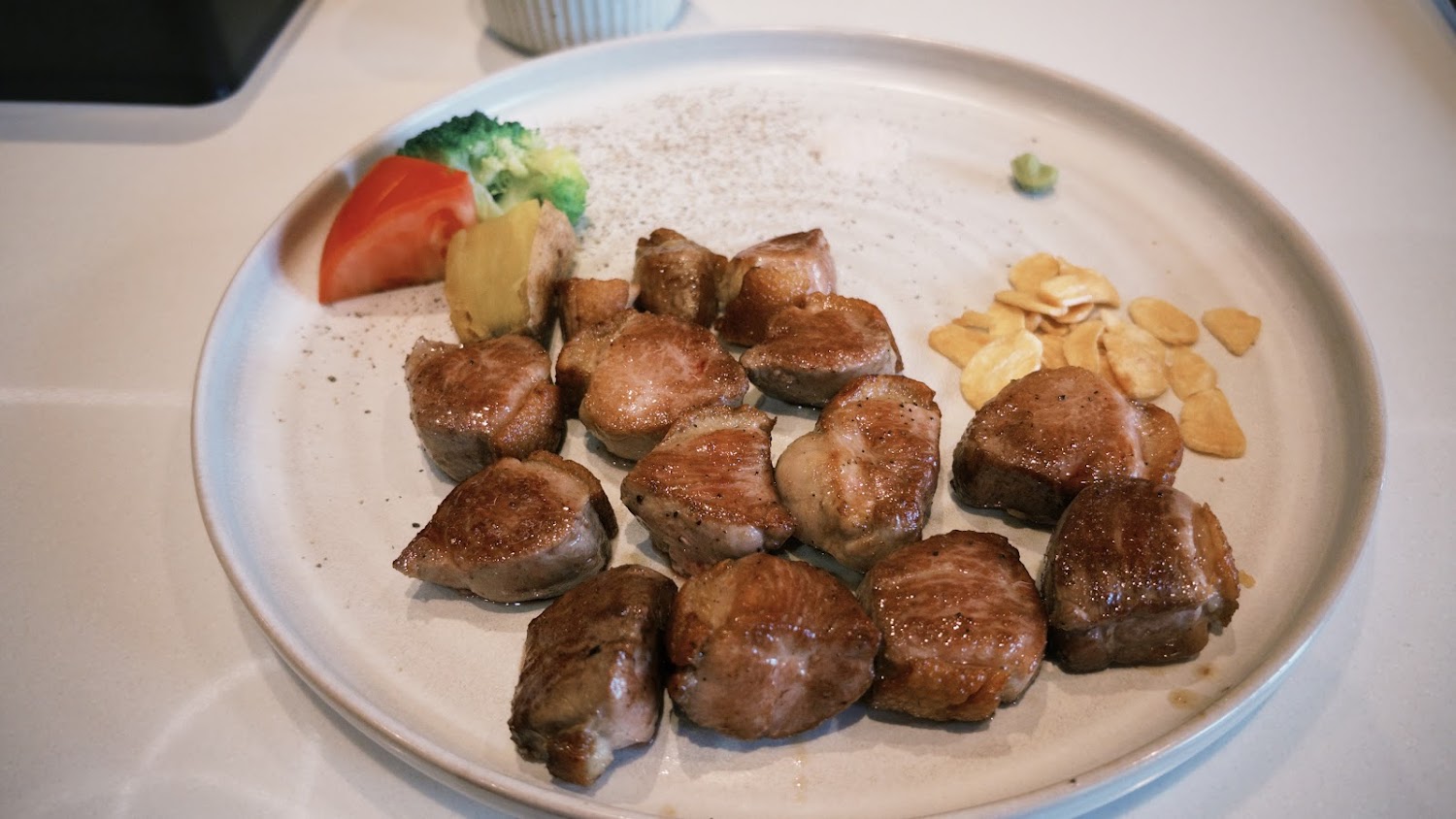 這張圖片主要是一盤煎牛肉塊，旁邊有配菜如番茄、花椰菜、薑片以及蒜片，並附有些調味料如胡椒鹽和芥末。