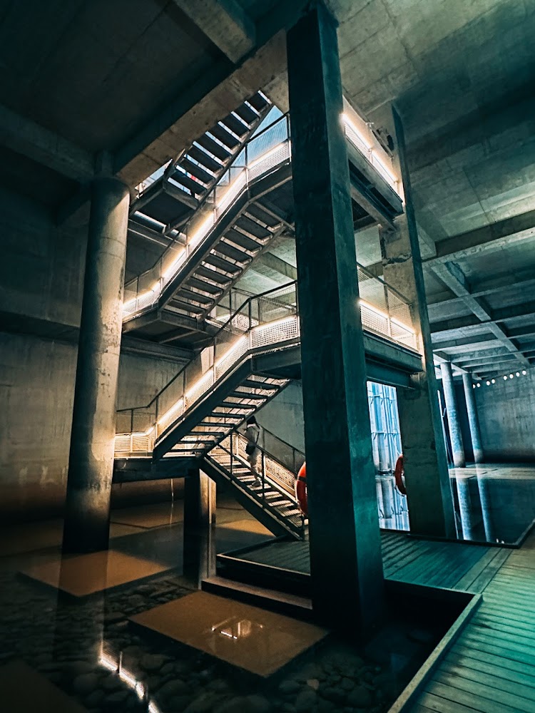 現代工業風格的混凝土建築內部，有光條裝飾的金屬樓梯，周圍有水面和木質棧道。