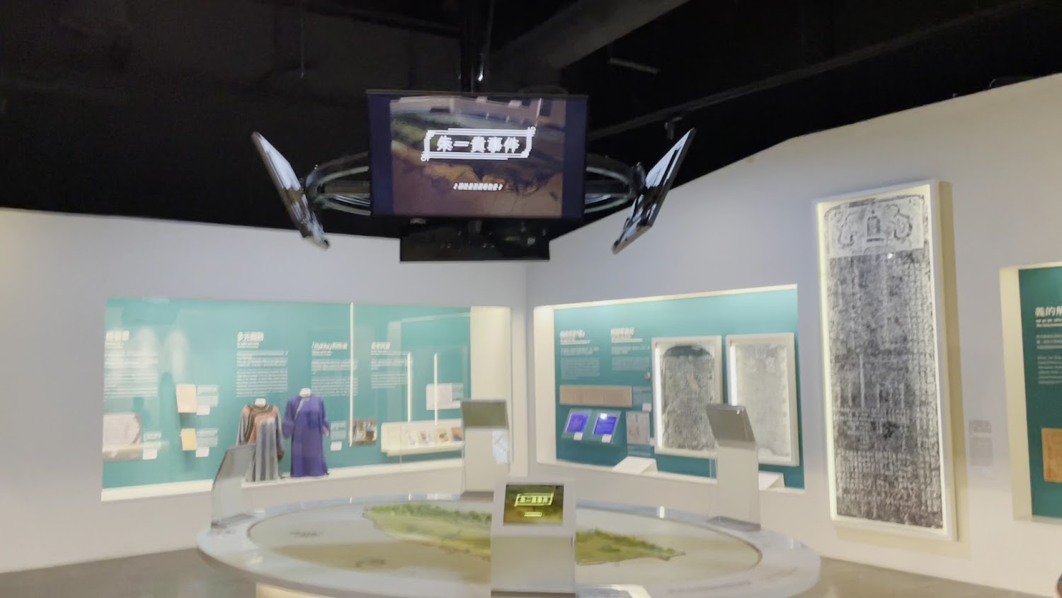 這張圖片展示了一個博物館展區，中心有地形立體模型，周圍有展示文物、古籍、石碑拓本和說明文字，並設有互動螢幕與懸掛的多媒體播放設備，內容豐富，富有歷史教育意義。