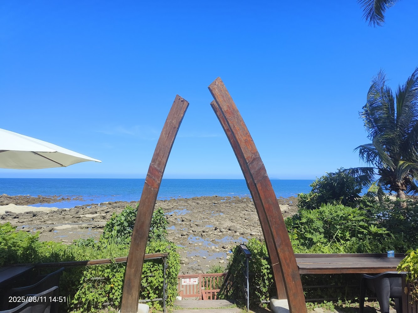 海邊景色，有兩根木頭豎立成拱門造型。前方是石灘與湛藍大海，兩側有椰子樹及綠色植物