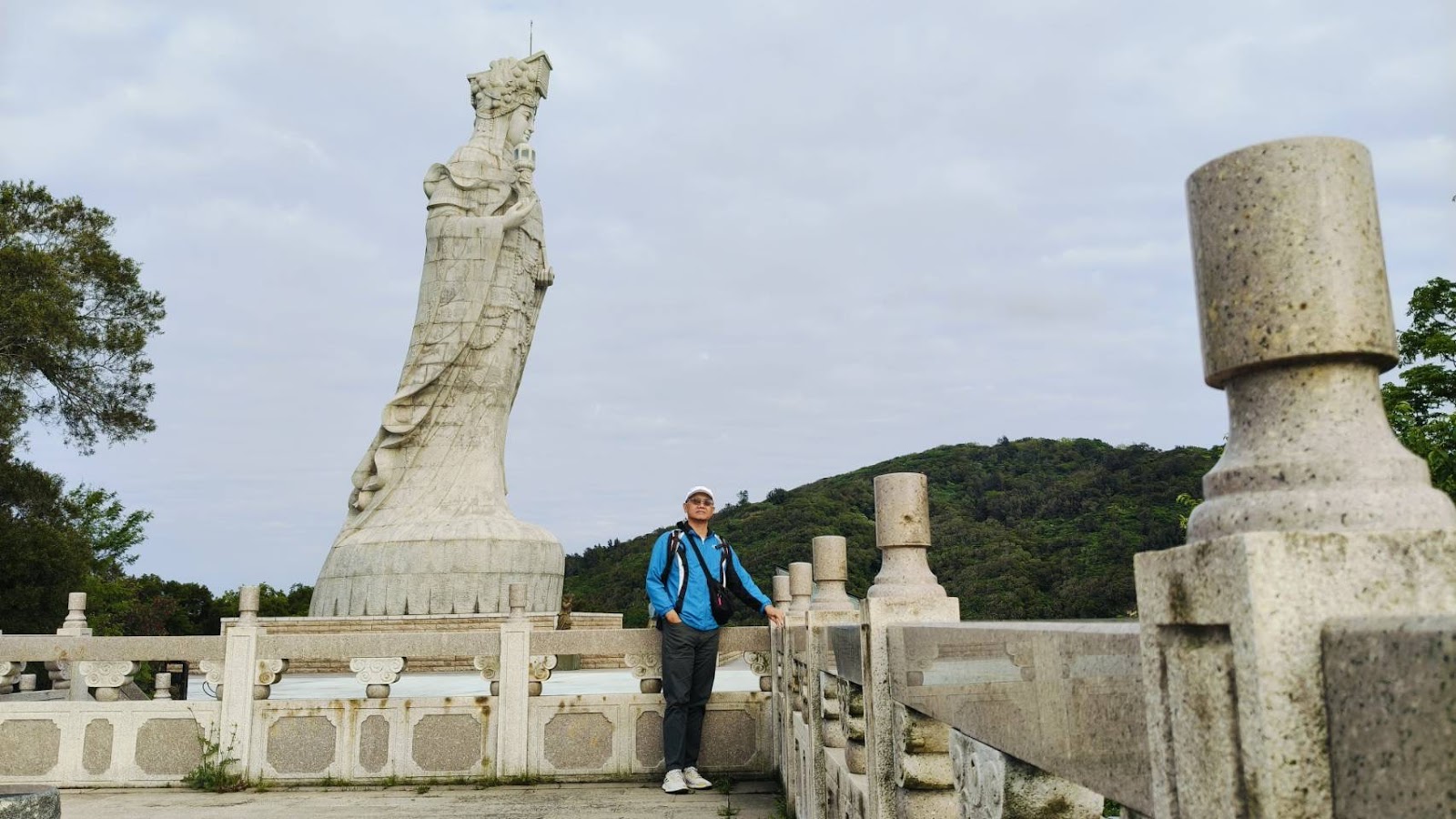 媽祖巨神像側面近景，遊客在石造欄杆旁仰望神像，陰天
