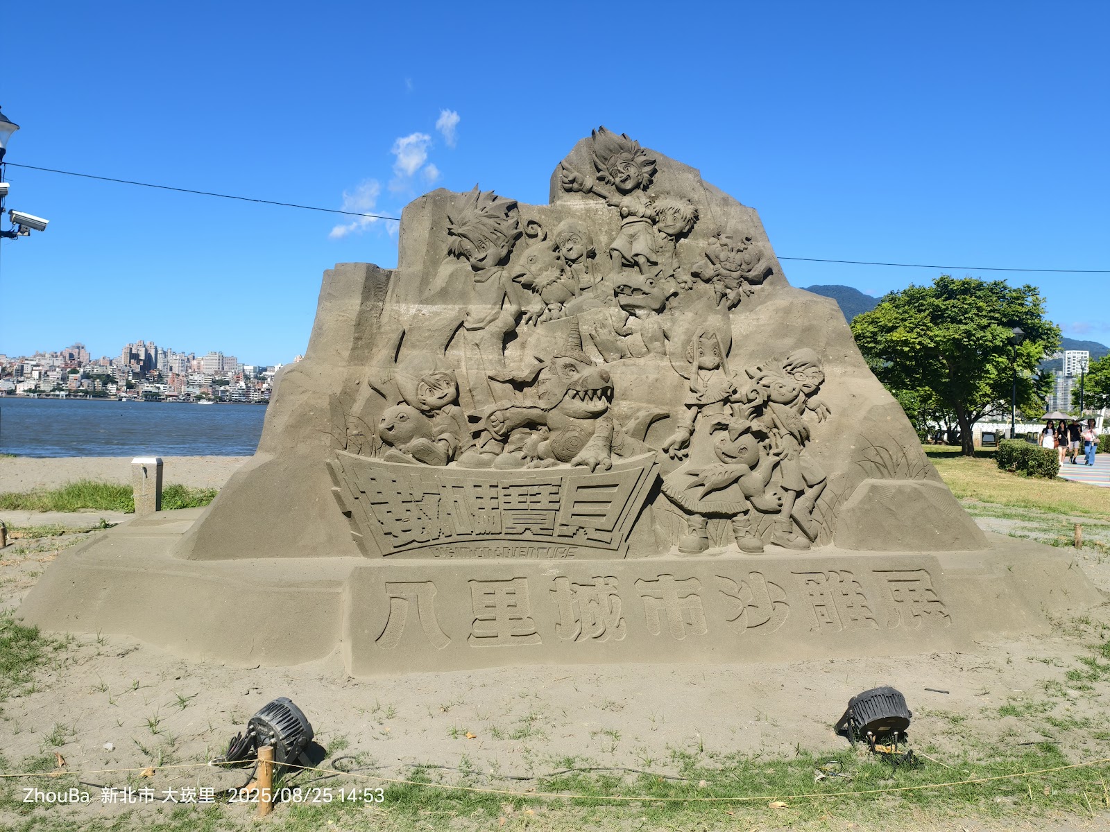 八里城市砂雕展，多款卡通角色砂雕群像，刻有八里城市沙雕展字樣，背景淡水河