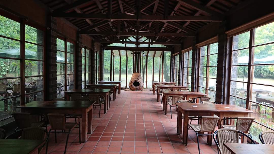 這張圖片展示一間充滿木質與自然氛圍的餐廳空間，擺放著多組木桌椅，兩側是大片落地窗，盡覽室外綠意，盡頭有一座磚砌壁爐，整體環境寧靜舒適。