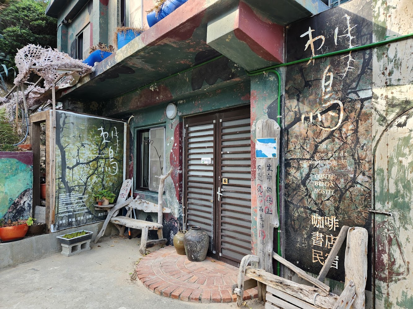 這是一家外觀充滿藝術氣息的咖啡書店民宿，門口有手工木椅、陶瓷花盆和綠色植栽，牆上寫著『咖啡 書屋 民宿』，環境溫馨且富有文藝氛圍。