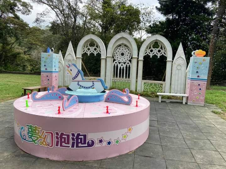 這張圖片展示了一個以童話風格設計的戶外裝置，有粉紅色圓台、可愛的搖馬與馬車裝飾，背景是仿哥德式拱門和草地，營造夢幻童趣的氛圍。