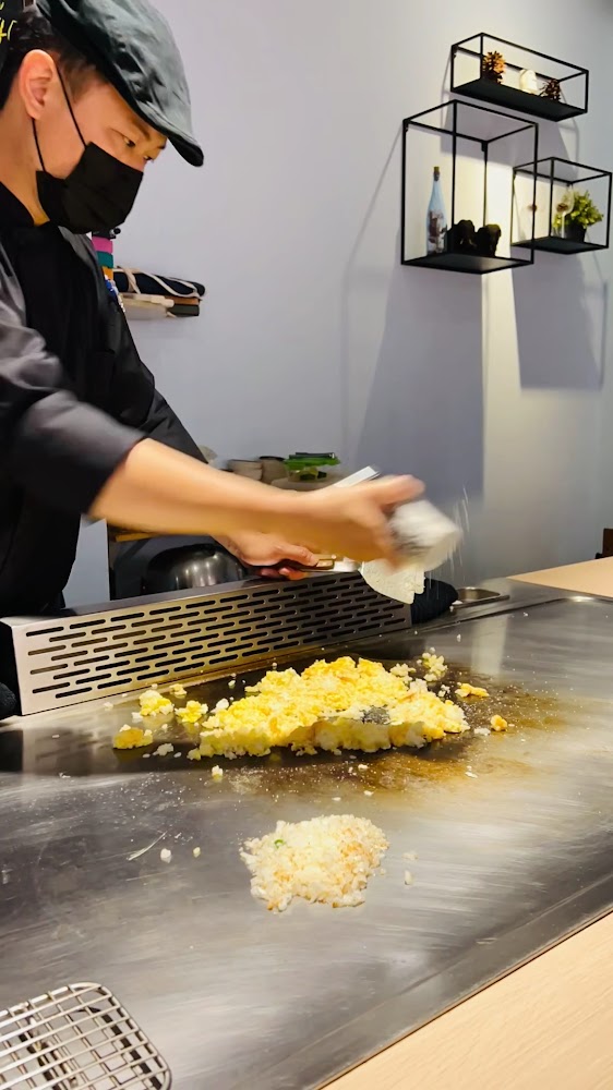這張圖片主要是一位廚師正在鐵板上料理炒飯，旁邊有調味料撒在食材上，背景則是簡約的牆上裝飾和擺設。