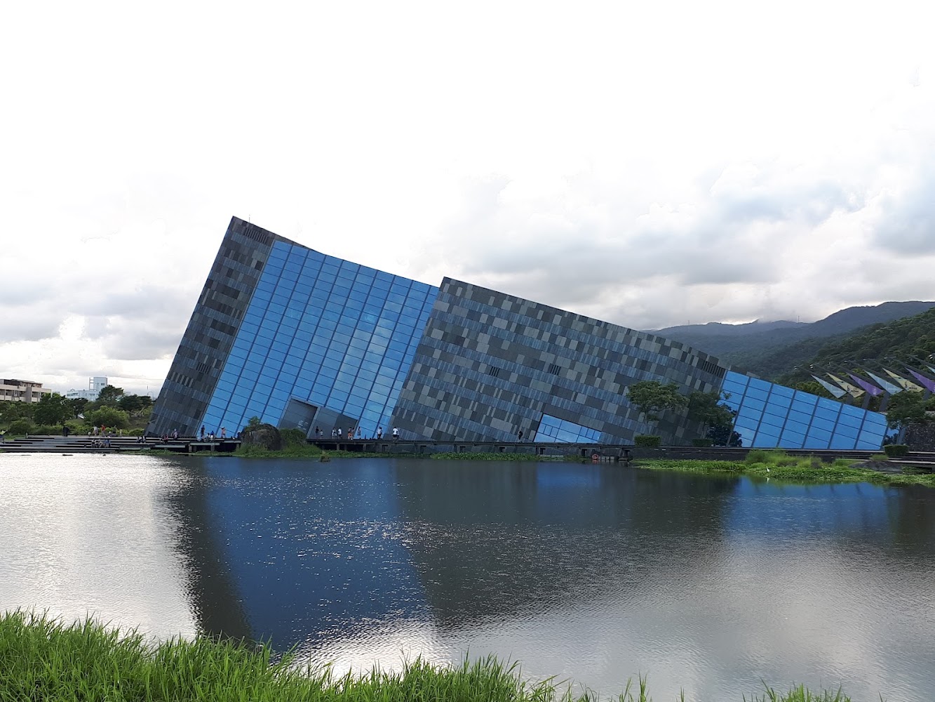 外觀傾斜且充滿現代感的建築坐落在湖邊，建築倒映在水面上，四周有綠地和山脈