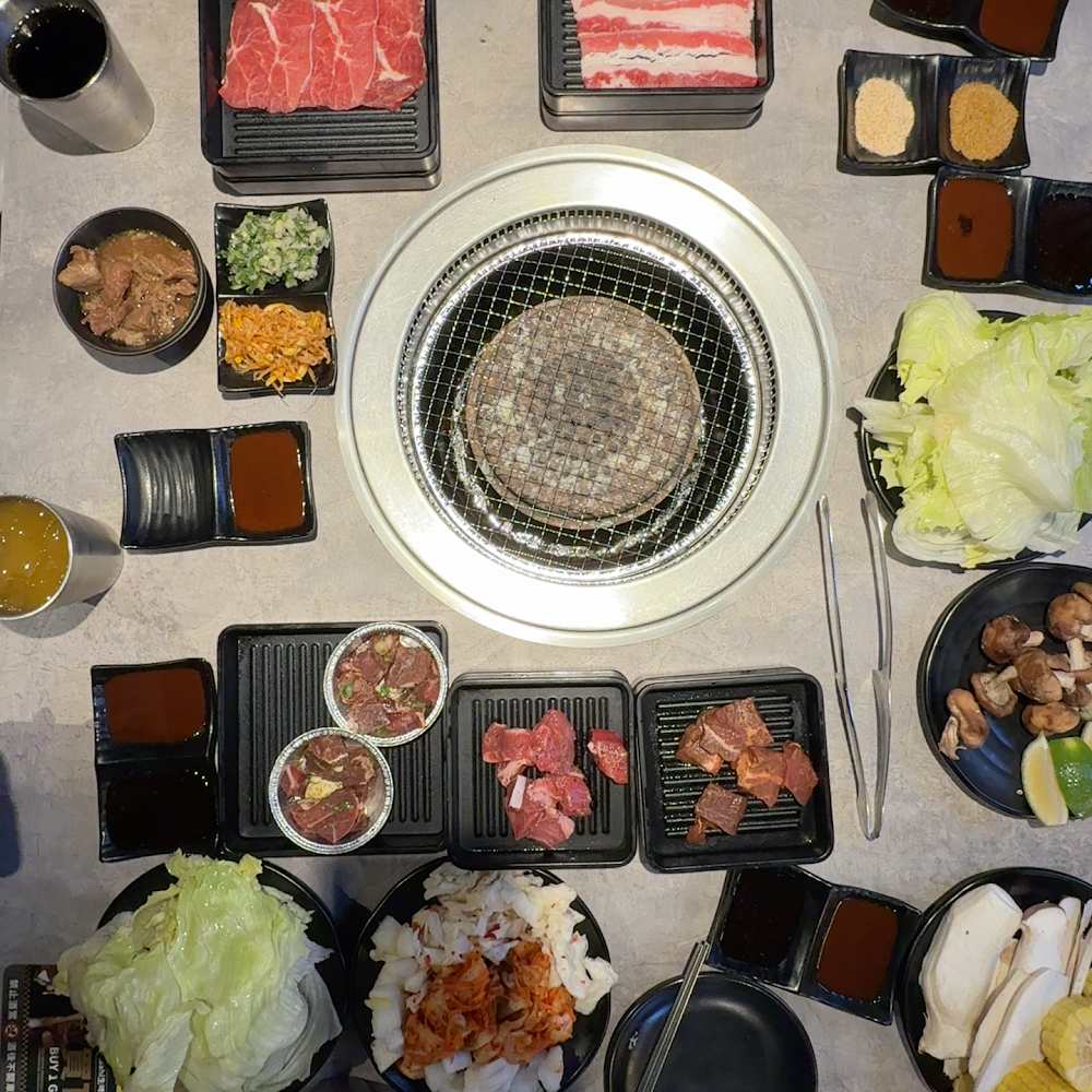 日式燒肉餐桌，有烤爐、各種生牛肉片、蔬菜、蘑菇、泡菜、生菜葉和多種醬料