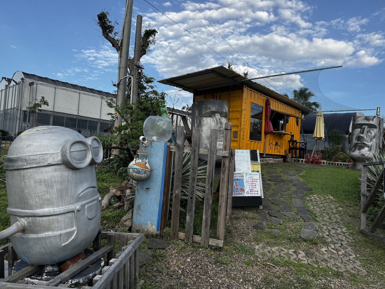 黃色貨櫃屋咖啡店，前方有小徑、告示牌與摩艾石像裝置藝術