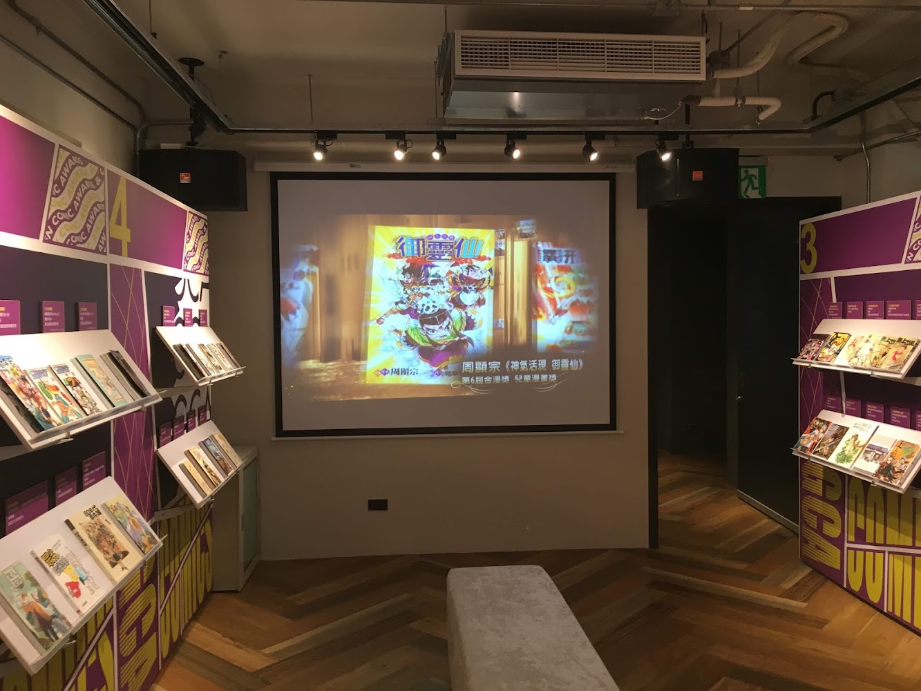 這張圖片顯示一間展示空間,兩側牆面整齊擺放多本漫畫,正前方有大螢幕播放漫畫相關影像,地板為木質拼花設計,整體空間明亮且帶有現代感。圖片無明顯浮水印。