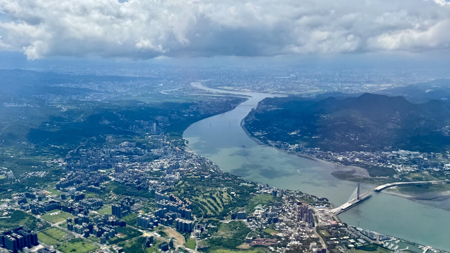 這張圖片俯瞰一條大型河流穿越城市與山區，河上有大橋，城市高樓與綠地交錯，遠方能見到天際線和雲層，展現自然與都市的融合景觀。