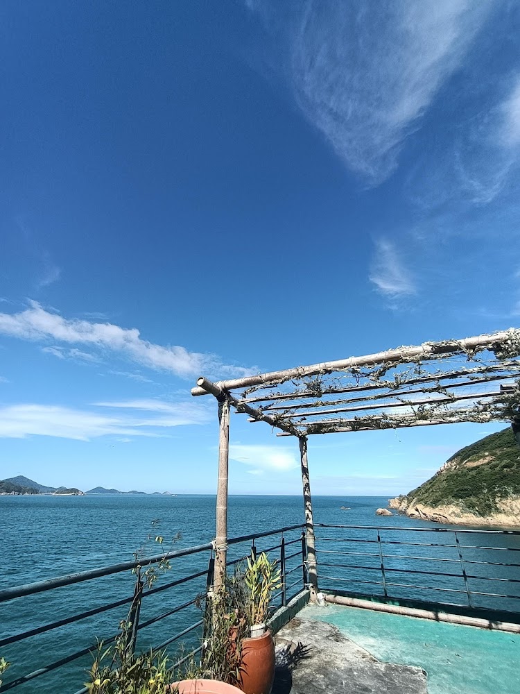 這張圖片展示一個臨海陽台，有竹製棚架與幾盆植物，旁邊是蔚藍大海與山丘，天氣晴朗，天空有美麗的雲朵，環境悠閒宜人。
