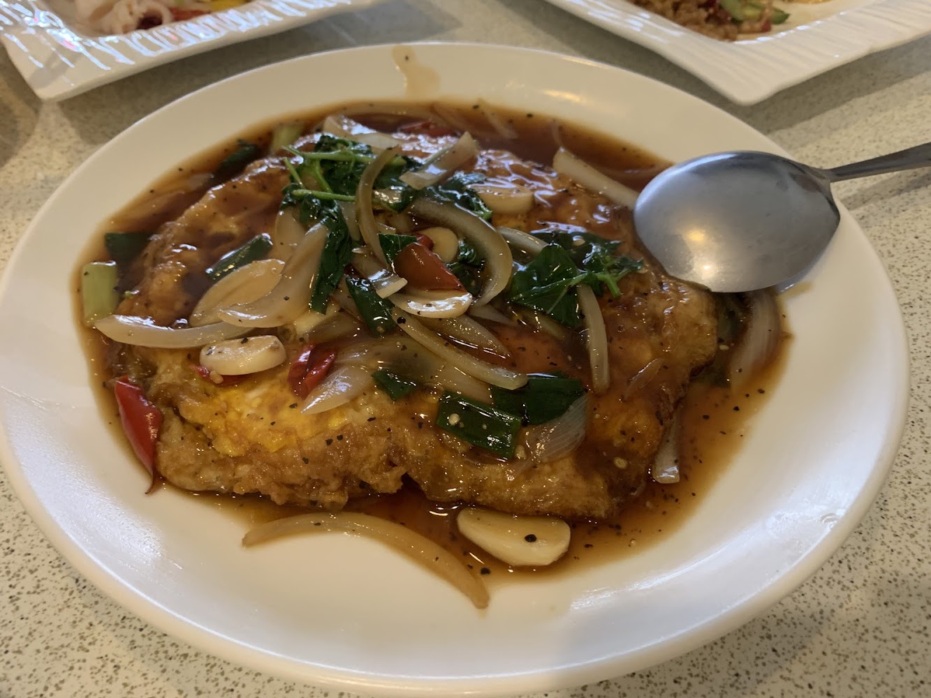 這張圖片主要是一道中式蛋料理，上面鋪有洋蔥、辣椒、蔥段及蒜片，再淋上濃郁的醬汁，看起來色香味俱全，旁邊配有一把湯匙。