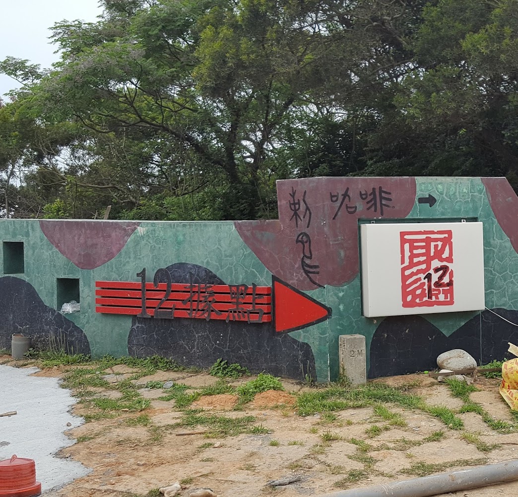 這張圖片主要是展示一處名為『12據點』的軍事設施入口，牆上有紅色指示及標誌牌，背景為樹林，牆面有迷彩塗裝，並帶有導向箭頭。