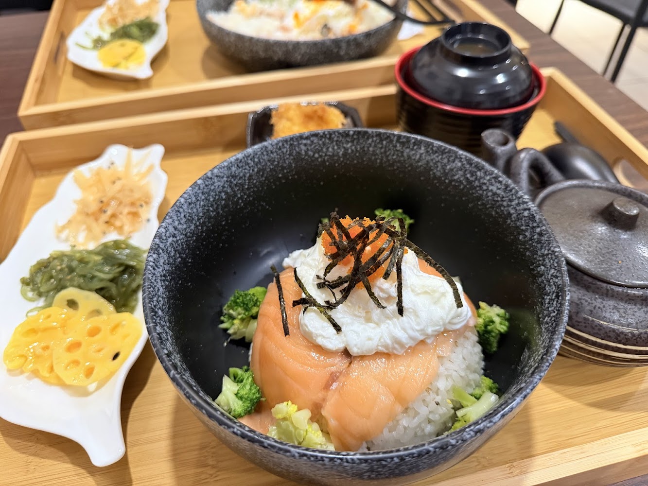 這張圖片展示了一份精緻的日式餐點，包括主菜鮭魚蓋飯，配有溏心蛋、蔬菜、海苔絲和魚卵，旁邊有三樣小菜以及湯品和茶具，擺盤美觀豐富。