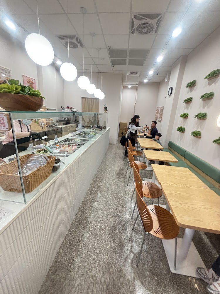 這張圖片是一間明亮現代的餐廳，設有開放式料理檯、木質餐桌椅、牆壁裝飾著蔬菜，氣氛溫馨，有顧客正在用餐。