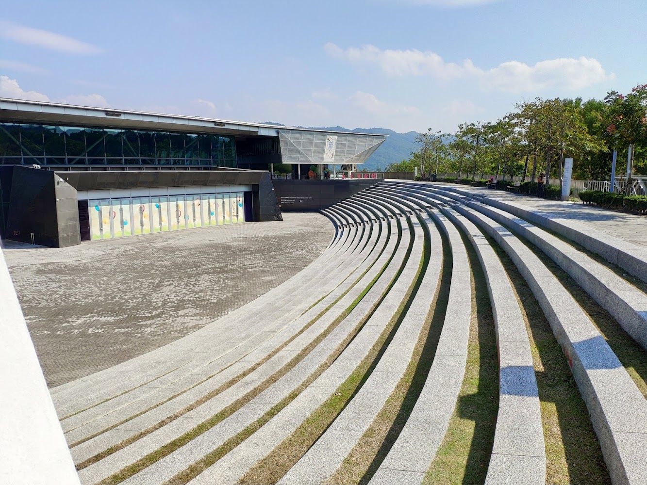 這張圖片主要展示了一個露天階梯式圓形廣場，前方有一棟現代感建築物，週圍環繞著樹木與藍天，充滿開放氛圍。