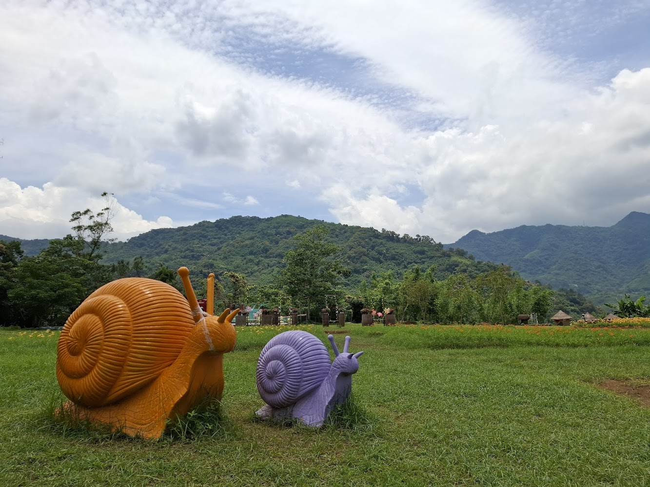 兩隻色彩鮮豔的巨型蝸牛雕塑位於草地上,背景是青山和藍天白雲