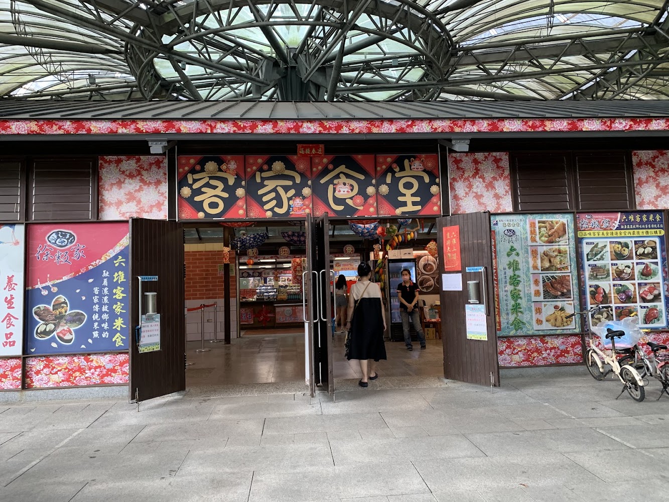 這是一間客家食堂，外觀有傳統花布裝飾，門口展示多樣客家料理的菜單，吸引顧客前來品嚐美食。