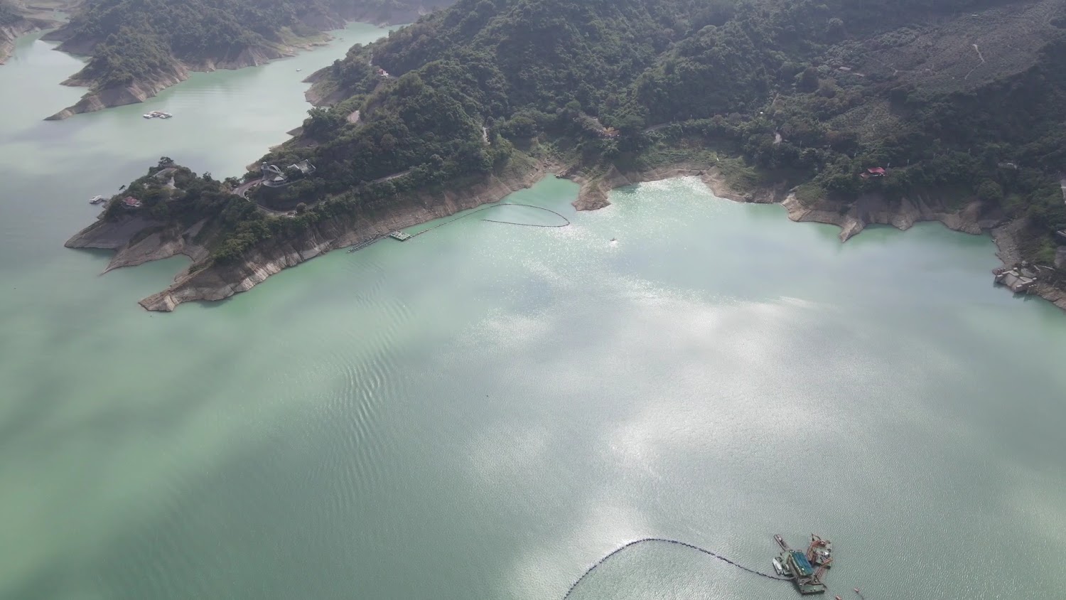 這張圖片主要顯示一座大型水庫,湖水碧綠,周圍環繞著山脈和林木。湖面上有幾座設施或船隻,岸邊可見山坡與部分建築。圖片未見明顯浮水印。