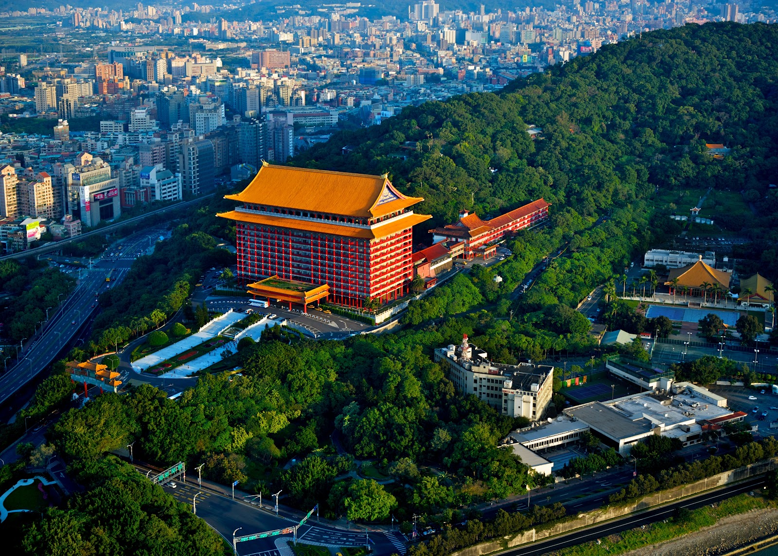 圓山大飯店鳥瞰全景，宮殿式紅色建築配金黃屋頂，雄踞綠色山頭，背景台北市區天際線