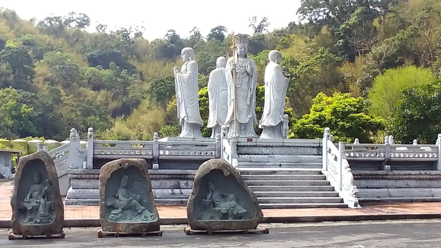 這張圖片主要展示了多尊佛教石雕像，包括前方三座嵌於石塊中的雕像，以及階梯上排列的四尊大型石雕，整體氛圍莊嚴寧靜，背景是茂密的樹林。