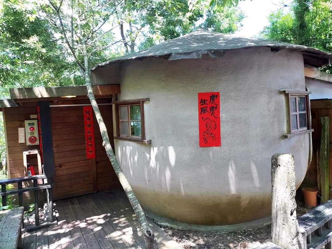 這張圖片主要是一間圓形土牆建築，外牆貼有紅色春聯，旁邊有木造結構與窗戶，周圍環繞著樹木和自然環境，展現樸實的鄉村風格。