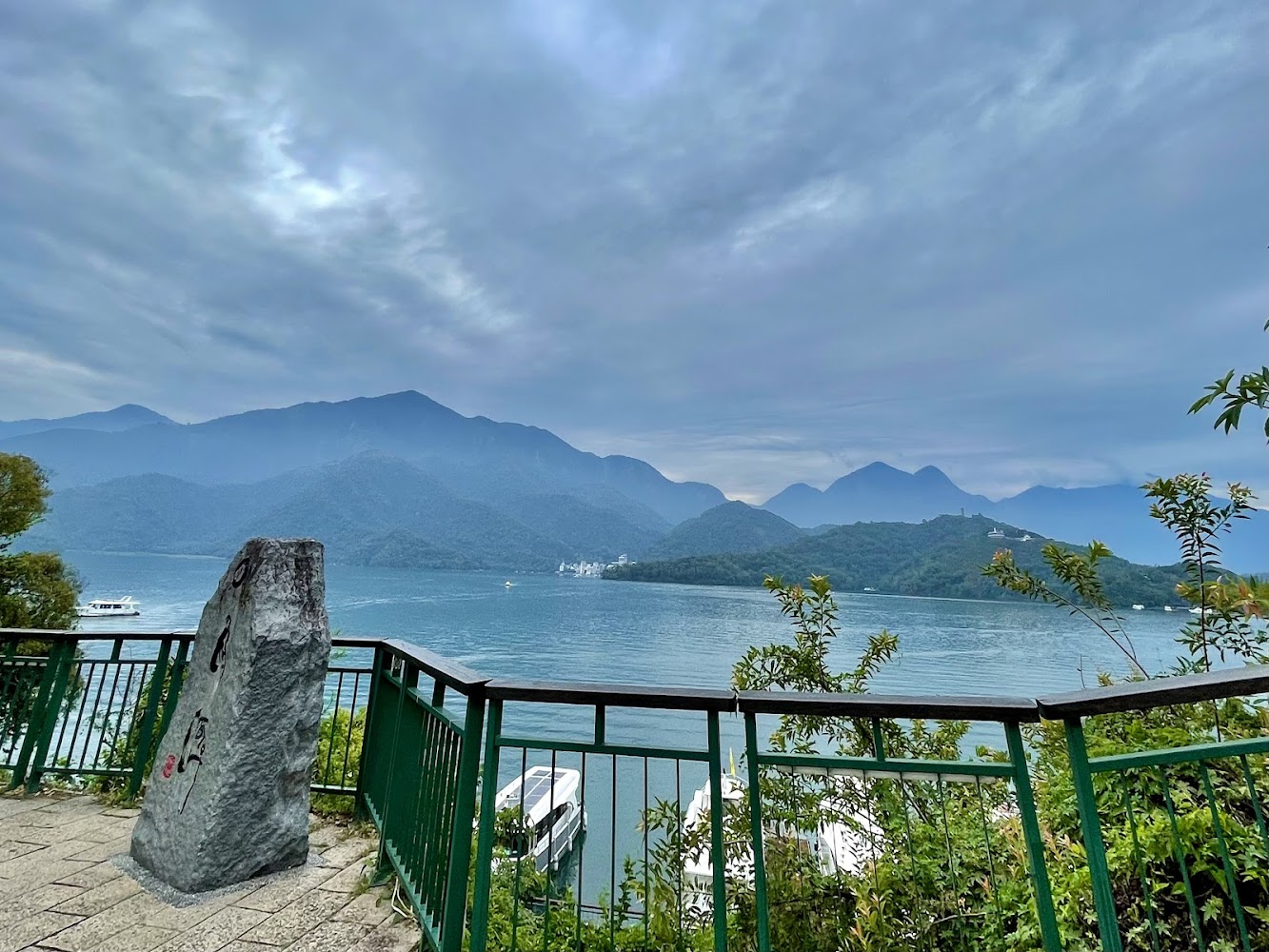 湖景觀景台，有欄杆、石碑與翠綠山水，遠處可見群山與湖泊，還有船隻航行其間