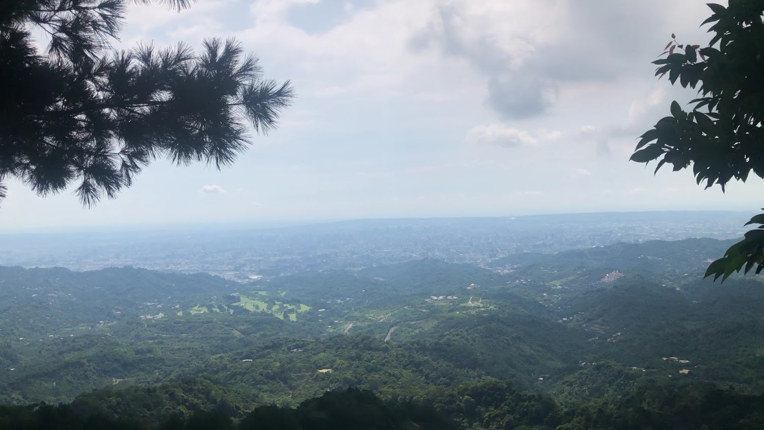 這張圖片主要展示了遼闊的山巒綠意景色，遠方隱約可見城市，天空中有雲朵飄浮，畫面邊緣有樹枝與葉子增添自然氛圍。