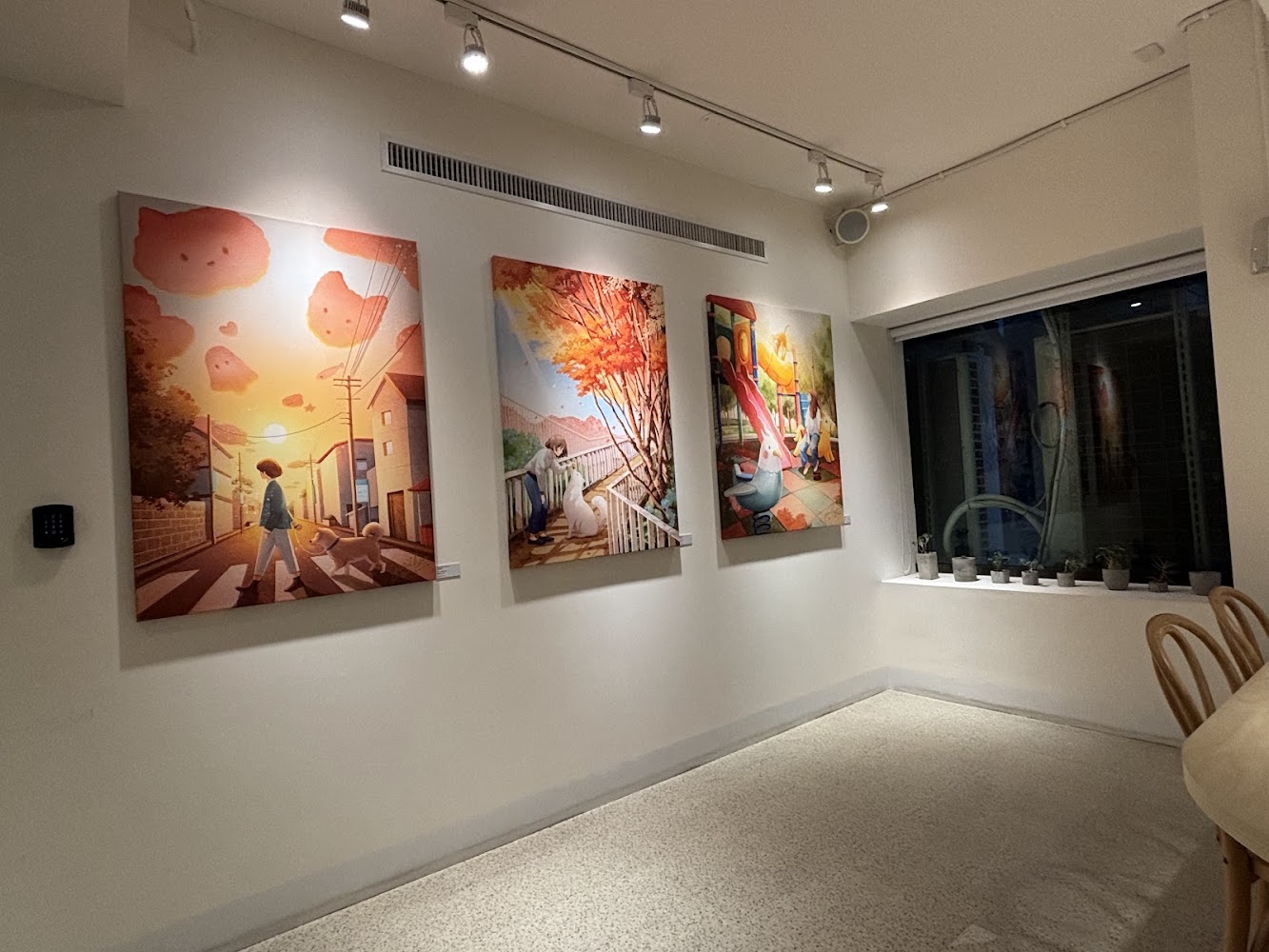這張圖片展示了一面牆,牆上掛有三幅色彩鮮明的插畫作品,內容包括人物、動物與自然景色。右側窗台上擺放數盆小盆栽,整個空間氛圍溫馨藝術感十足。