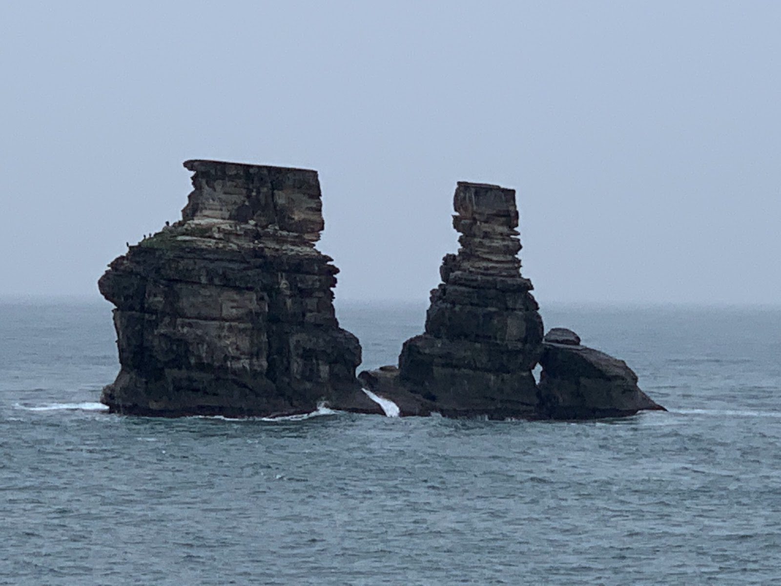 兩座層疊砂岩礁岩聳立灰色海面，陰天霧灰