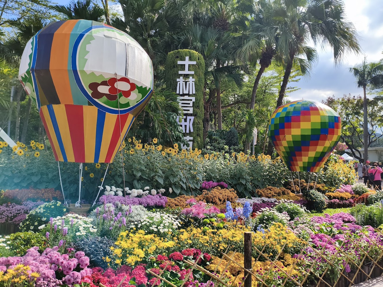 兩個彩色熱氣球裝飾與周圍盛開的繽紛花卉，高大棕櫚樹與白色中文招牌相襯