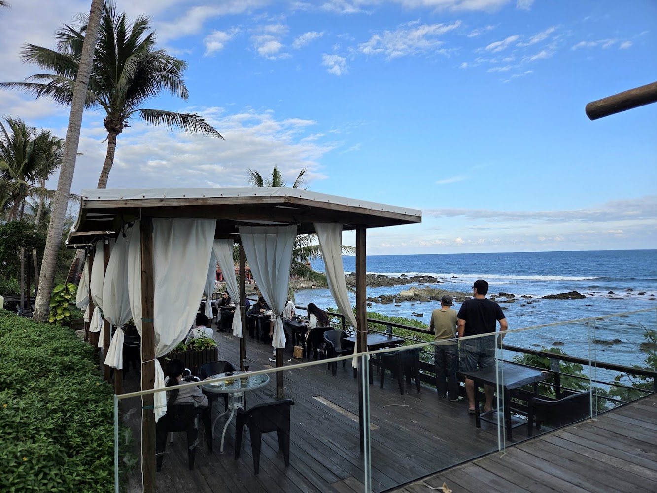 海邊露天咖啡廳，有遮陽棚和白色窗簾，旅客坐在椅子上享受美景