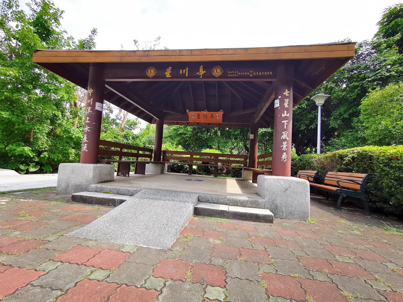 木造涼亭周圍被綠樹與灌木環繞，旁邊有公園長椅