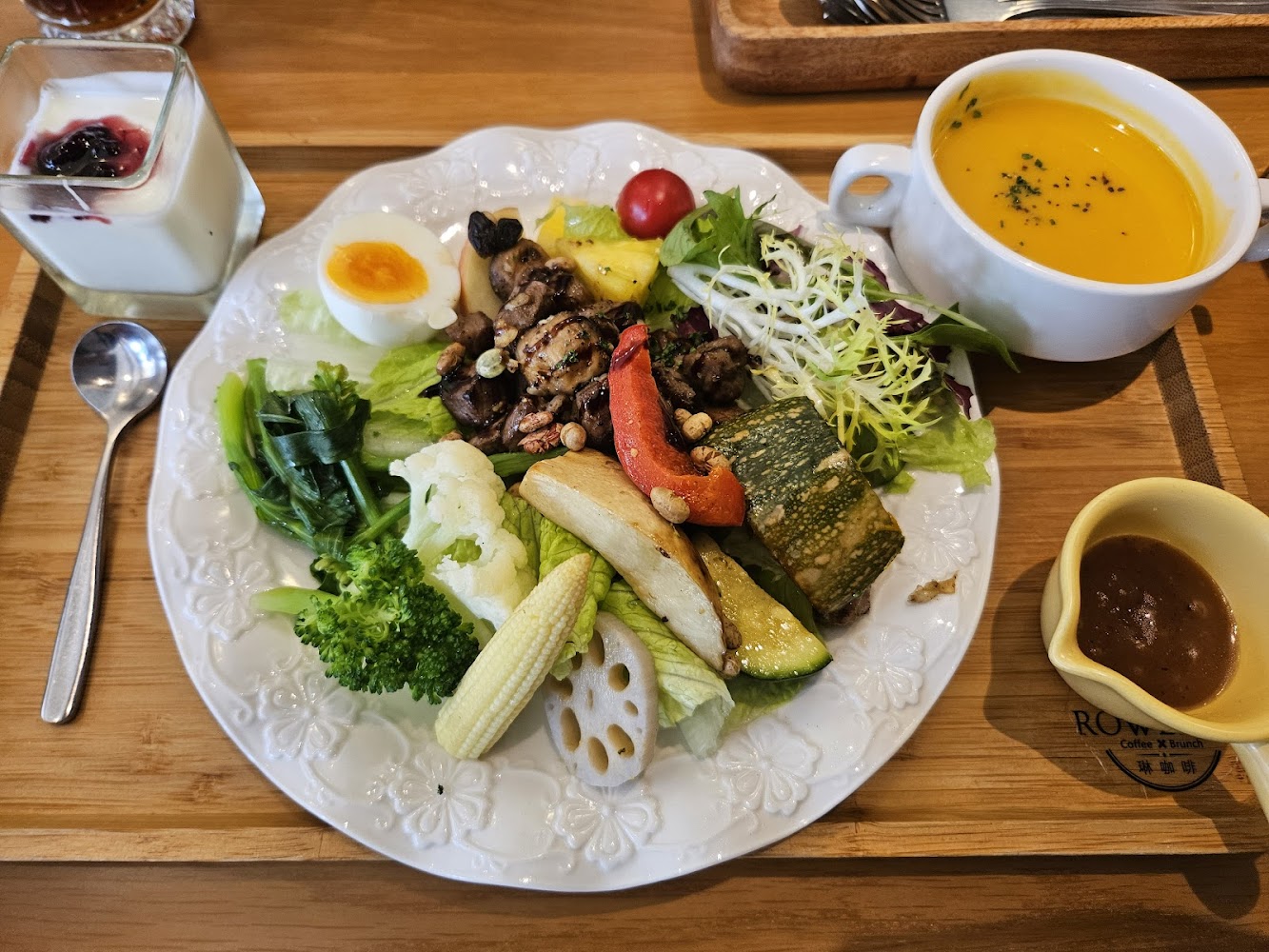 這張圖片是一份豐富的早餐套餐，包含多種新鮮蔬菜、半熟蛋、烤蘑菇、水果、生菜沙拉及蘸醬，旁邊有一碗南瓜濃湯與優格甜點，營養均衡。