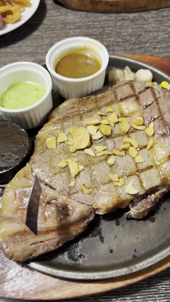 鐵板牛排上有烤痕與蒜片，旁邊附兩種醬料與烤石。