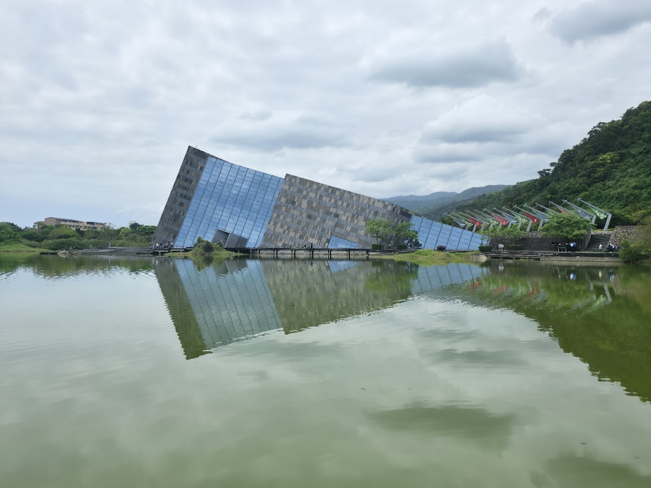 傾斜造型建築矗立湖畔，湖面倒映著建築和天空，遠處有山林與旗幟裝飾