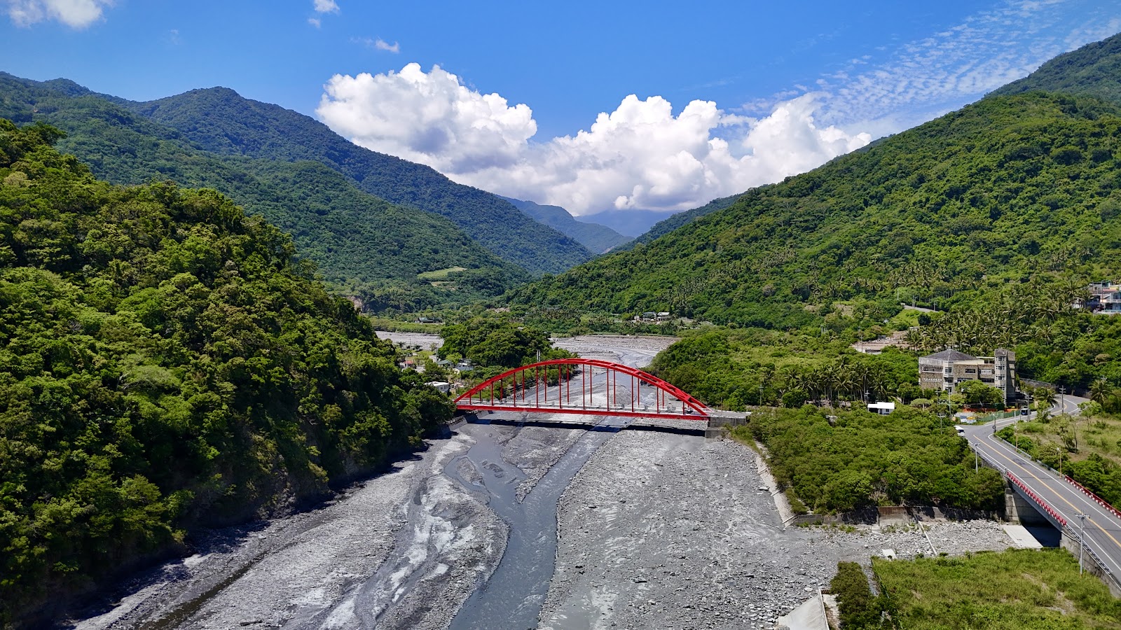 航拍俯視金崙溫泉虹橋全景,紅色拱橋橫跨溪床,兩側青山環繞,藍天白雲,右側公路延伸