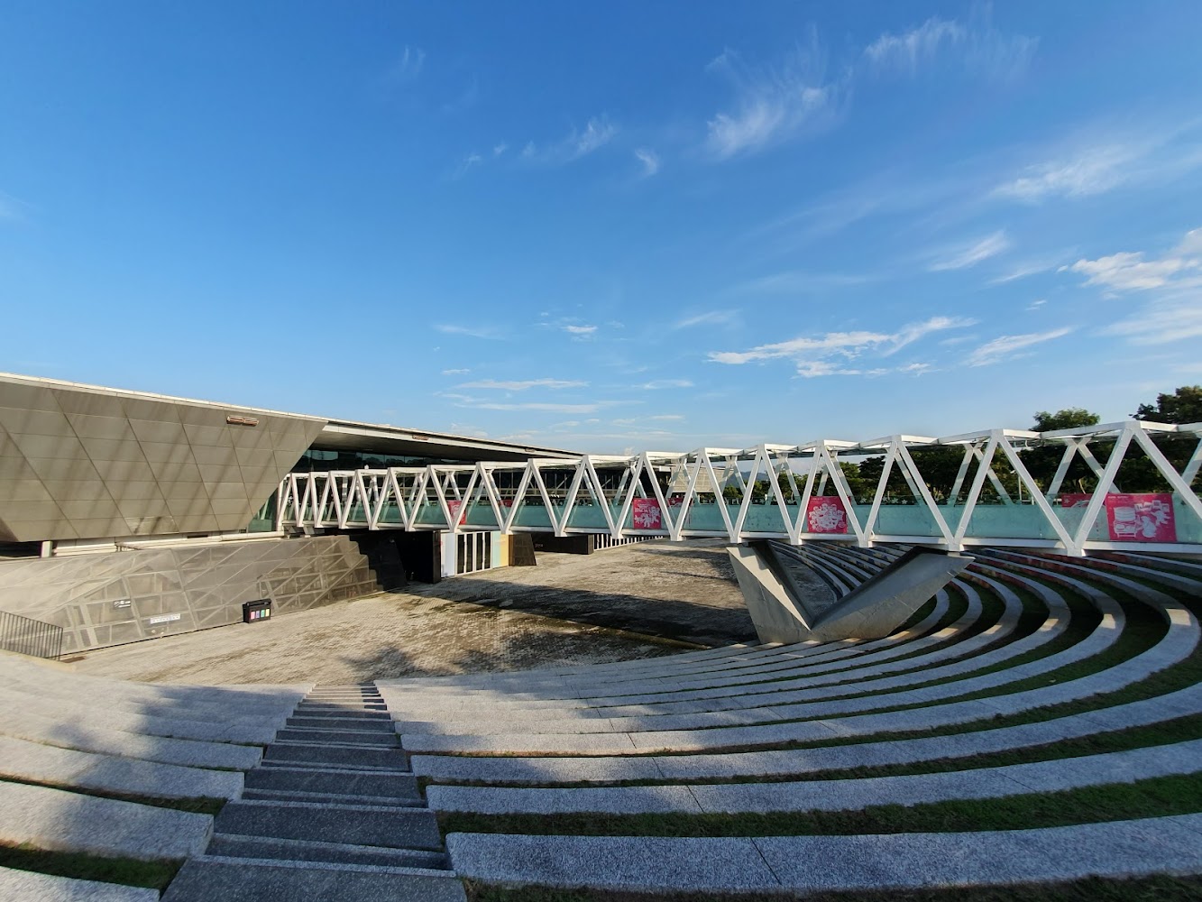 圖片中有一座現代化建築，右側是一條白色鋼構天橋，橋下是圓形階梯廣場，整體設計充滿現代感，天氣晴朗，天空蔚藍。