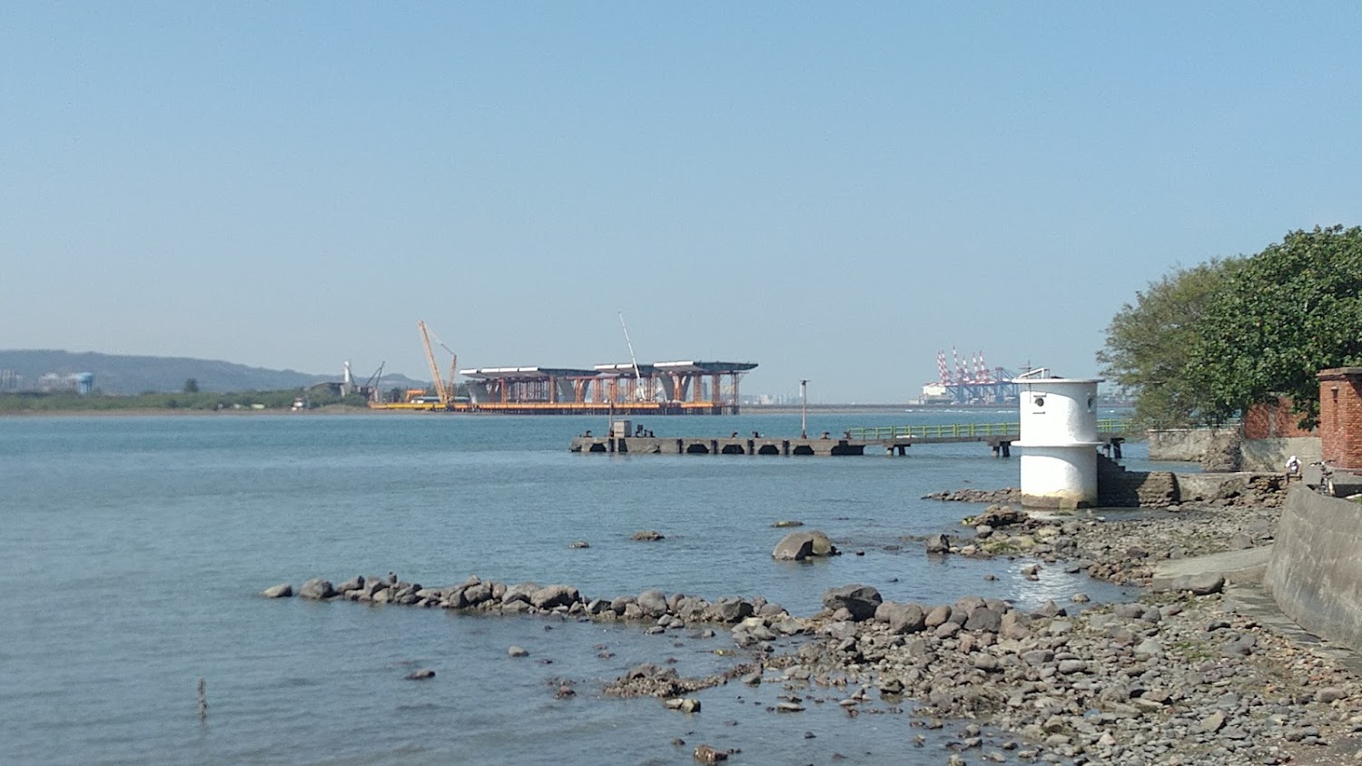 河岸景色前景有礫石灘與白色圓柱哨所,遠方可見施工橋樑與起重機