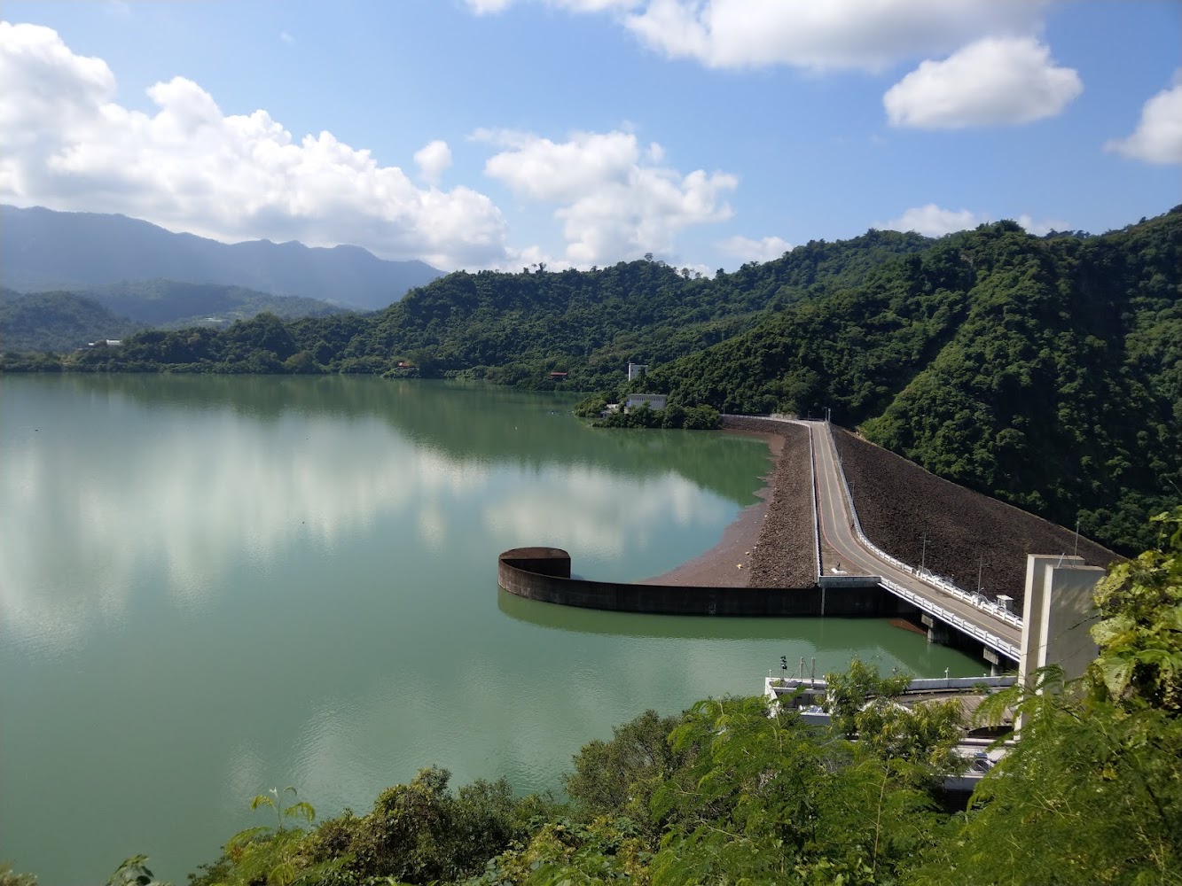 這張圖片主要呈現一座大型水庫,畫面中央是廣闊的湖面,右側有一座大壩和道路,四周環繞著青翠的山林,天氣晴朗。圖片中無明顯浮水印。