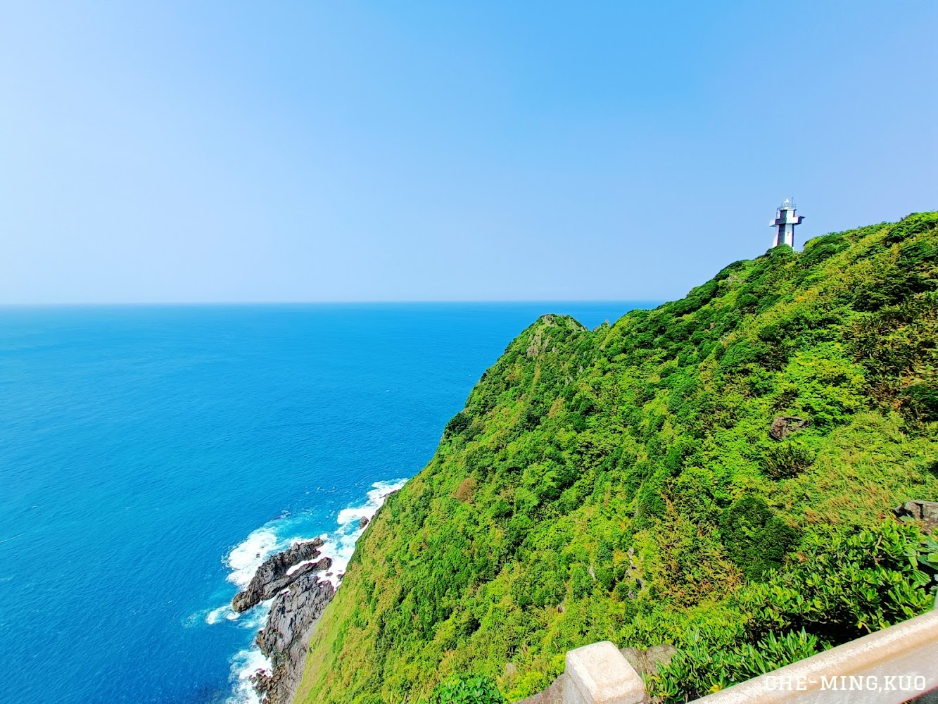 這張圖片主要展示蔚藍的海洋、綠意盎然的山坡，以及山頂上的一座燈塔，構成壯麗的自然與人文景觀。